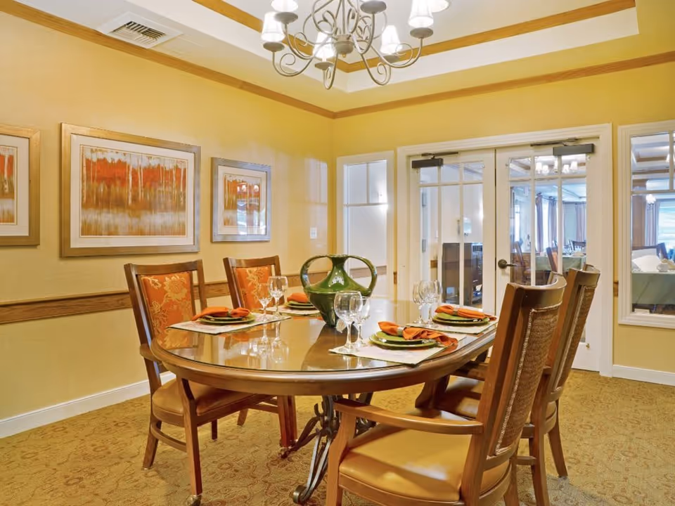 A dining room with a wooden oval table set for six people with green plates, orange napkins, and wine glasses. The room has yellow walls with three framed abstract paintings and a chandelier hanging from the ceiling. There are glass double doors leading to another room.