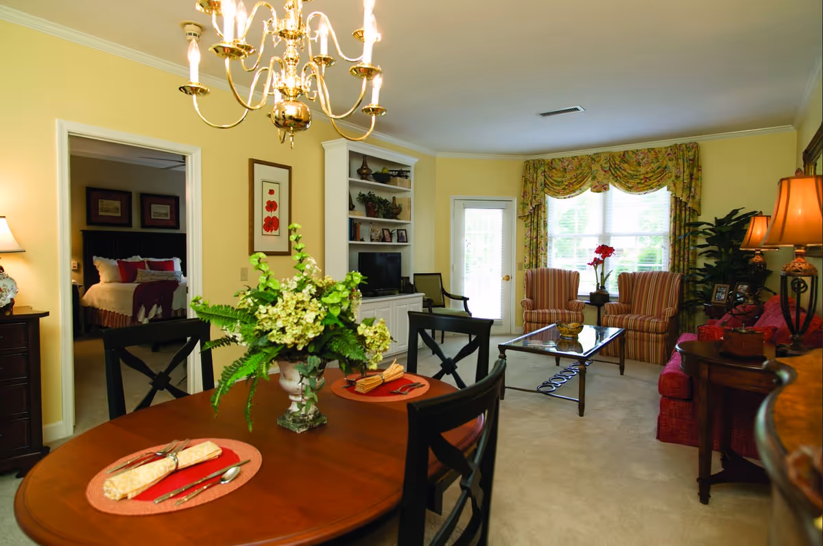 A cozy living and dining area in a retirement community with a wooden dining table set for two, a floral centerpiece, a chandelier above, and a living room with striped armchairs, a glass coffee table, a red sofa, and a large window with floral curtains. A bedroom with a bed and lamps is visible through an open doorway.