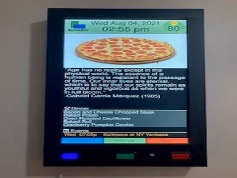 Digital display screen showing the date Wednesday August 4, 2021, time 2:55 pm, and temperature 80 degrees. The screen features an image of a pepperoni pizza, a quote about age by Gabriel Garcia Marquez, the dinner menu listing bacon and cheese chopped steak, baked potato, oven roasted cauliflower, baked roll, and cranberry pumpkin cookie, and an event listing for a baseball game between the Baltimore Orioles and New York Yankees.