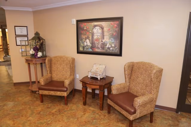 A cozy seating area in a rehabilitation and healthcare center featuring two patterned armchairs with brown leather seats, a small wooden side table between them holding a decorative item, a framed floral and bicycle artwork on the beige wall, and a round wooden table with a birdcage and flower arrangement in the corner.