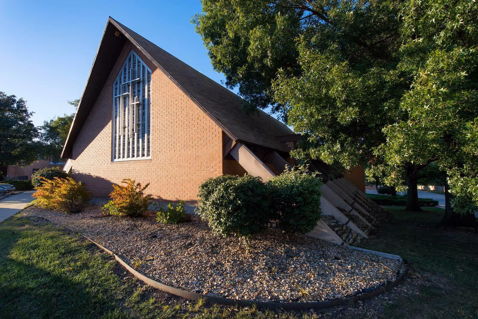 Exterior view of a brick building with a steeply pitched roof and a large vertical window featuring a cross design. The building is surrounded by landscaped bushes, trees, and a gravel bed under a clear blue sky.