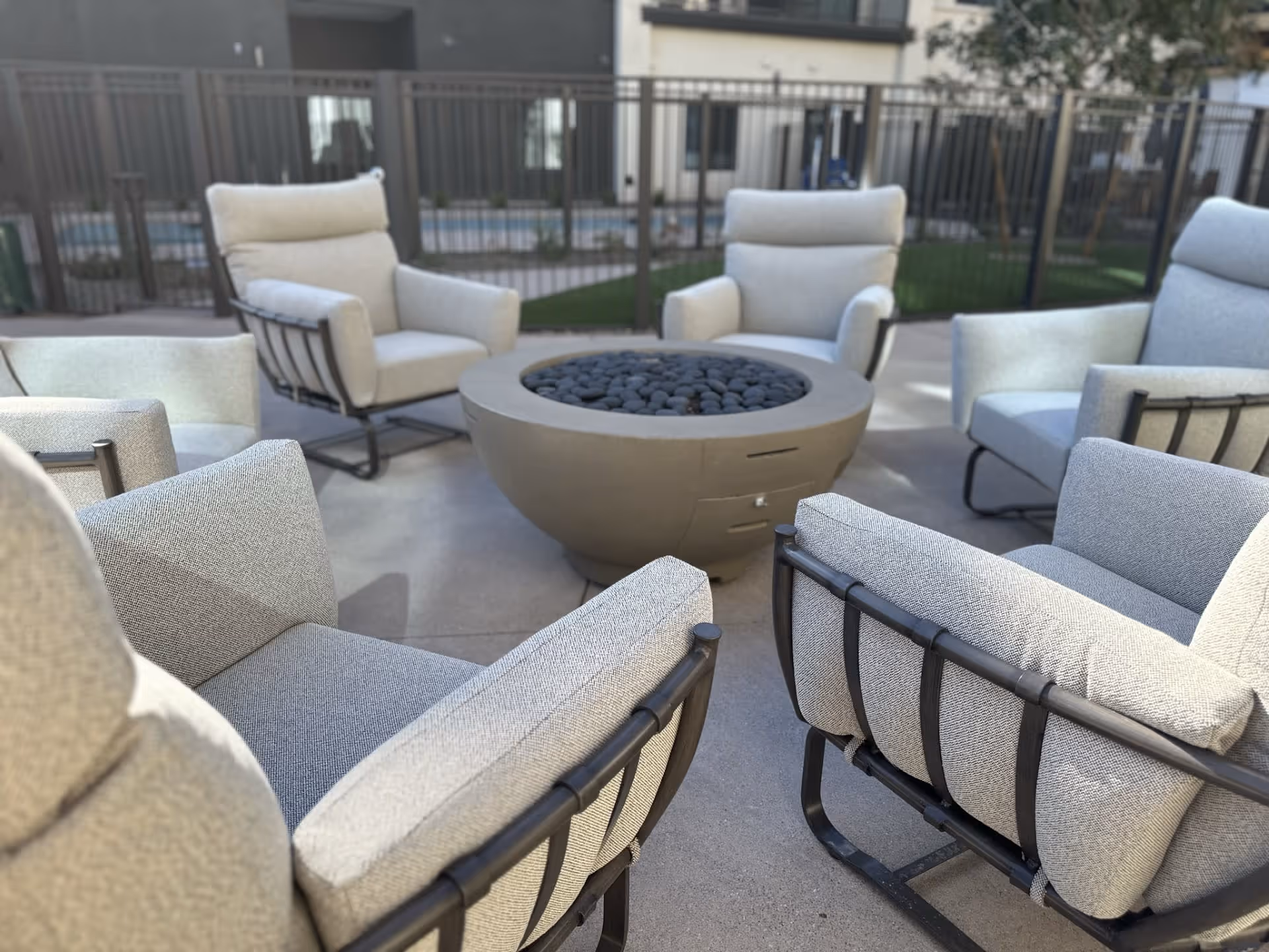Outdoor seating area with six cushioned armchairs arranged in a circle around a round fire pit filled with black stones, set on a concrete patio with a fenced background and some greenery.