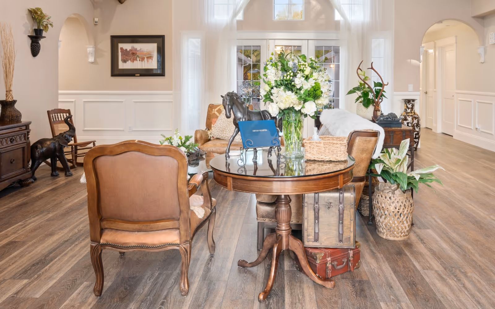 Bright, elegant living room seating area with a round wooden table, leather chairs, a large floral centerpiece, and hardwood floors.