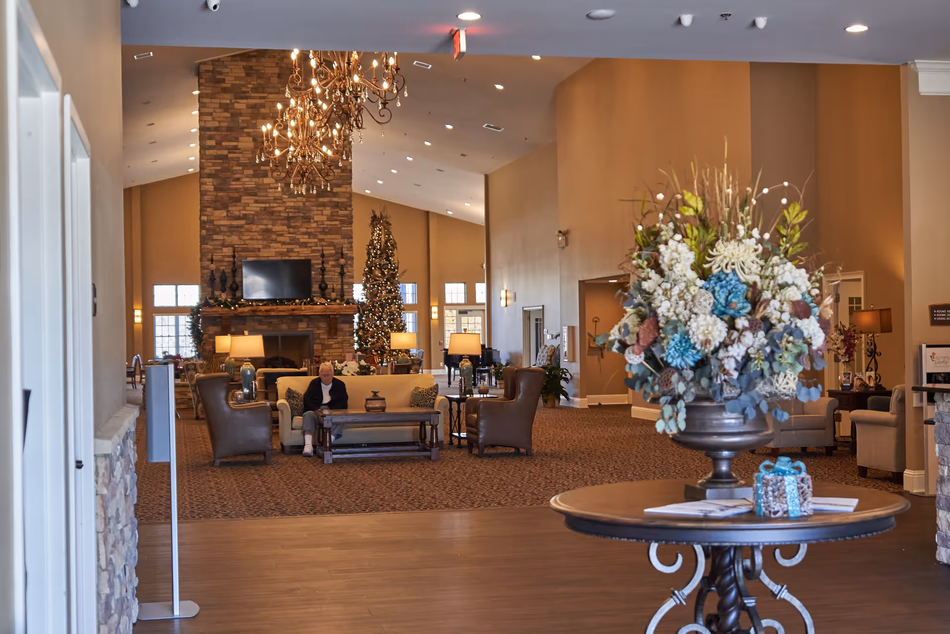 A spacious and warmly decorated senior living facility common area with a large stone fireplace, a Christmas tree, chandeliers, and comfortable seating including a sofa and armchairs. A person is sitting on the sofa. In the foreground, there is a round table with a large floral arrangement and a small decorative gift box.