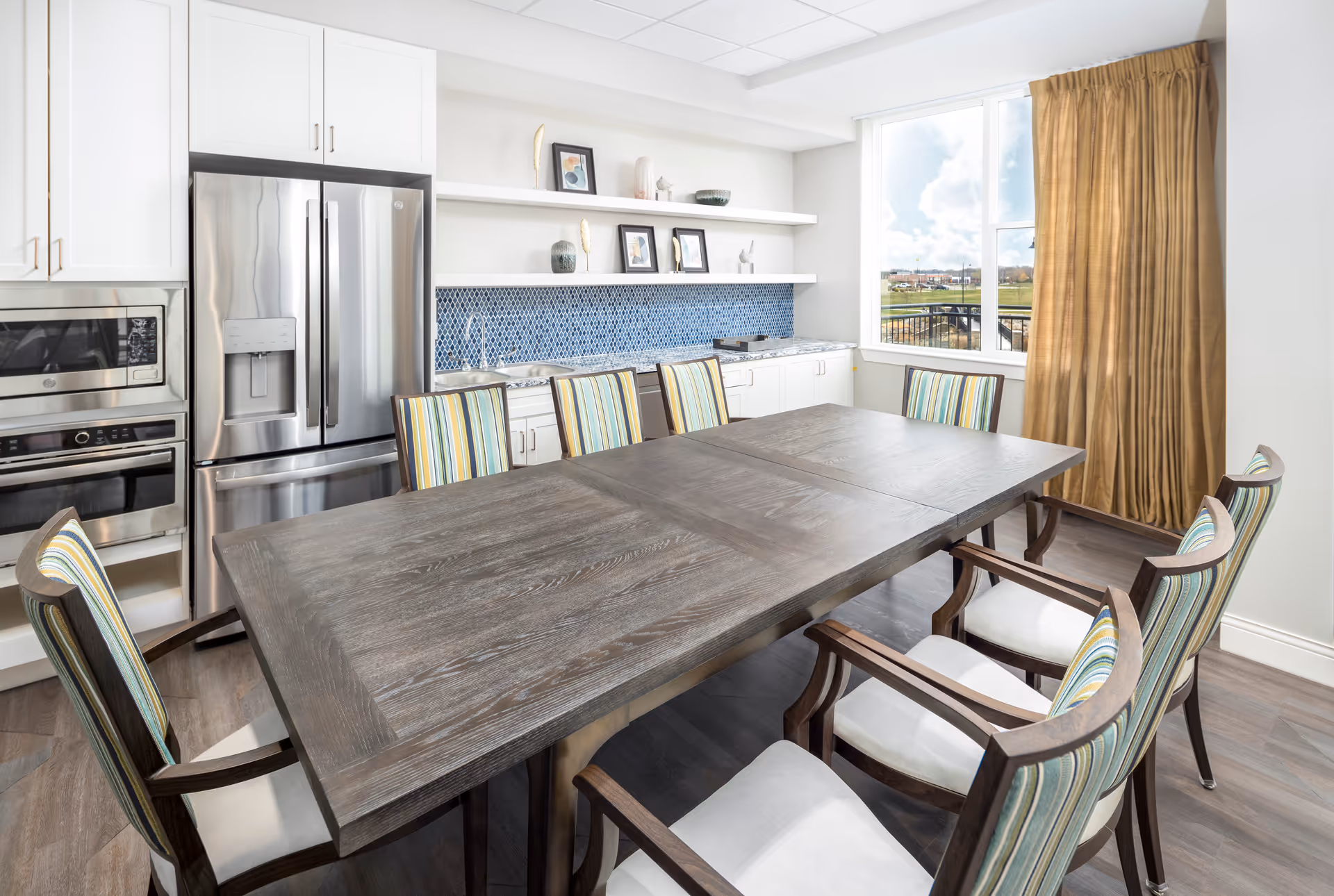 A bright dining area with a large wooden table surrounded by eight chairs with striped upholstery. The room features a modern kitchen area with white cabinets, a stainless steel refrigerator, built-in microwave and oven, and a blue tiled backsplash. A large window with gold curtains allows natural light to fill the space.
