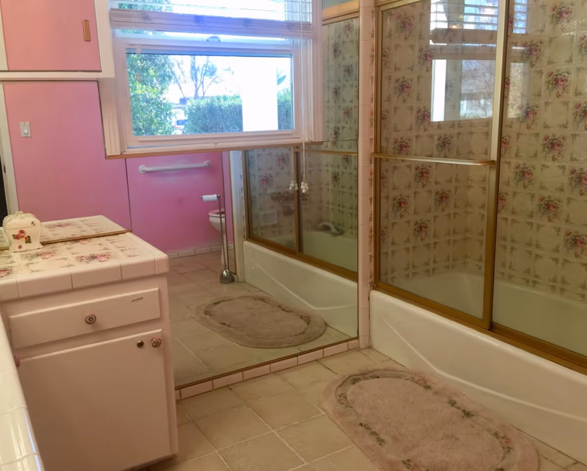 A bathroom with pink walls and floral patterned tiles in the shower area. The shower has sliding glass doors with gold trim. There is a window above the bathtub, a vanity with floral tile countertop and pink cabinetry, a toilet partially visible in the reflection of a large mirror, and two beige oval bath rugs on the tiled floor.