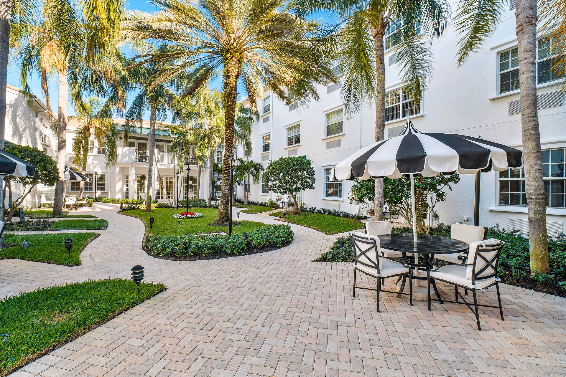 Sunlit courtyard with palm trees, paved walkways and a black-and-white umbrella shading a table and chairs in front of a white building.