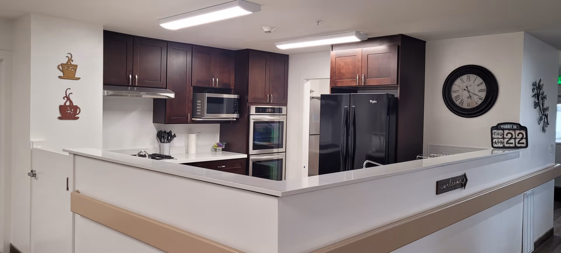 Modern kitchen area with dark wood cabinets, a black refrigerator, built-in microwave, double oven, and a white countertop. The kitchen is partially enclosed by a half wall with a beige handrail. Wall decorations include two coffee cup plaques, a round clock, and a calendar showing Thursday, April 22.