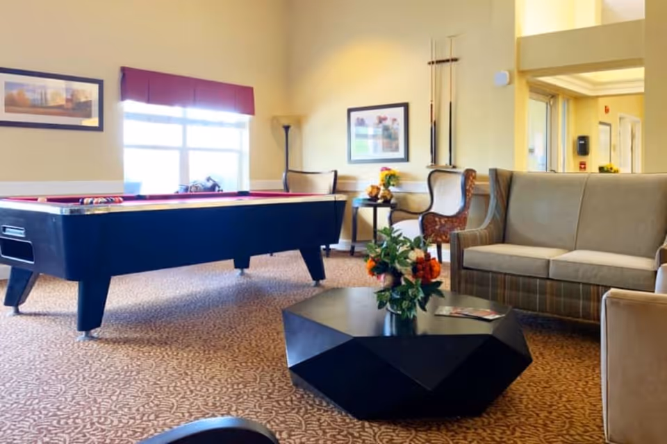 A bright and cozy common area featuring a pool table, a geometric black coffee table with a flower arrangement, a beige sofa, two armchairs, and framed artwork on the walls. The room has a large window with a red valance and carpeted flooring.