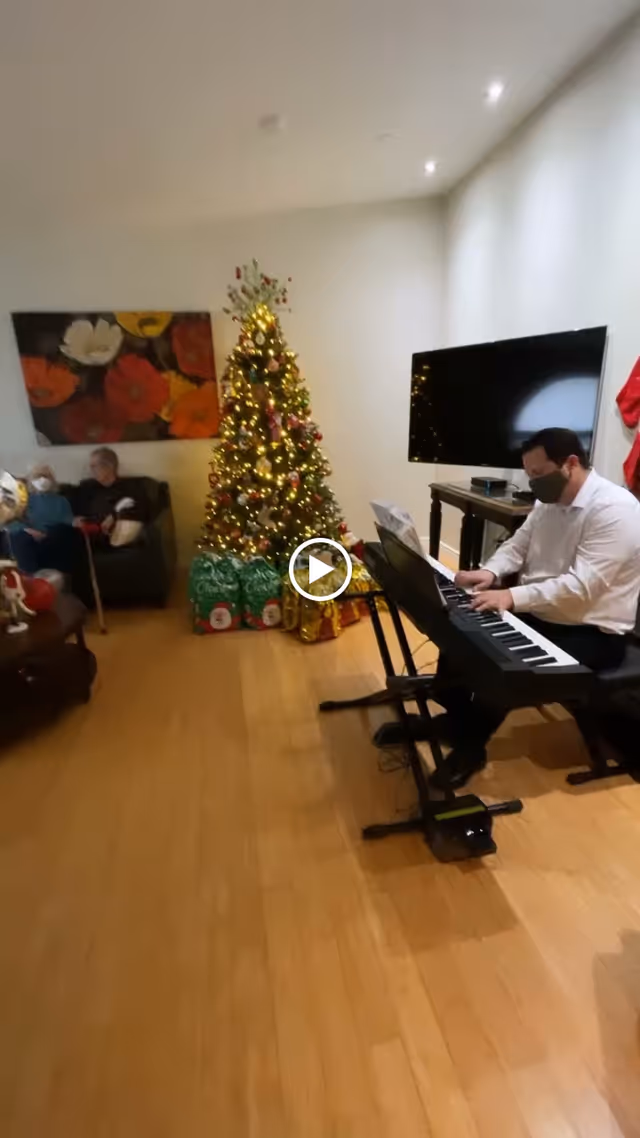 A man wearing a face mask plays an electric keyboard in a living room decorated for Christmas with a lit Christmas tree and wrapped presents. Two elderly people sit on a couch in the background near a colorful floral painting on the wall.