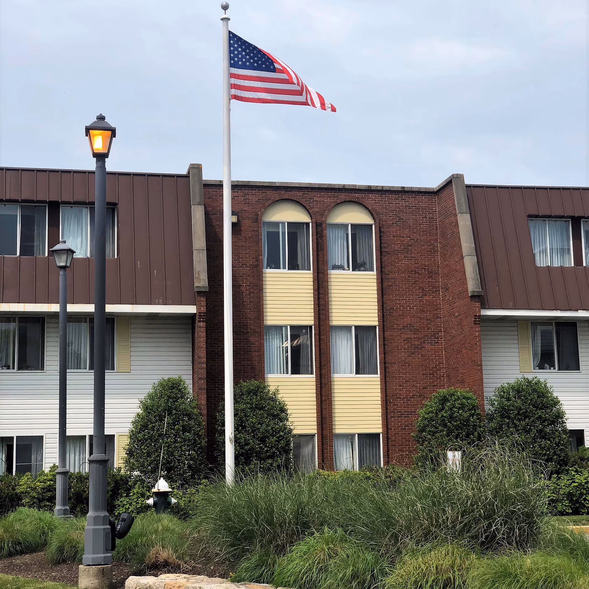 Exterior view of a multi-story brick and siding building with several windows, an American flag on a flagpole in front, two street lamps, and landscaped bushes and grass.