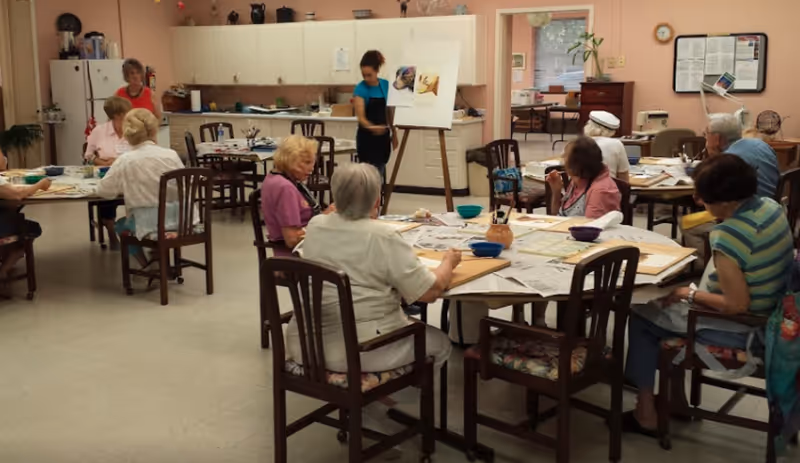 A group of elderly residents sit around tables in a communal activity room doing arts and crafts while a staff member stands near an easel.