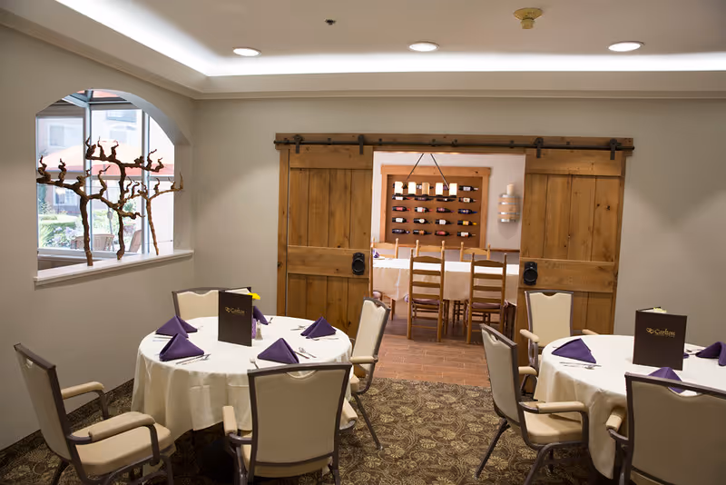 Dining area with round tables covered in white tablecloths, each set with purple folded napkins and menus. Beige cushioned chairs surround the tables. A window with a decorative branch arrangement is on the left wall. In the background, a wooden sliding barn door opens to another room with a long dining table and chairs, and a wine rack on the wall.