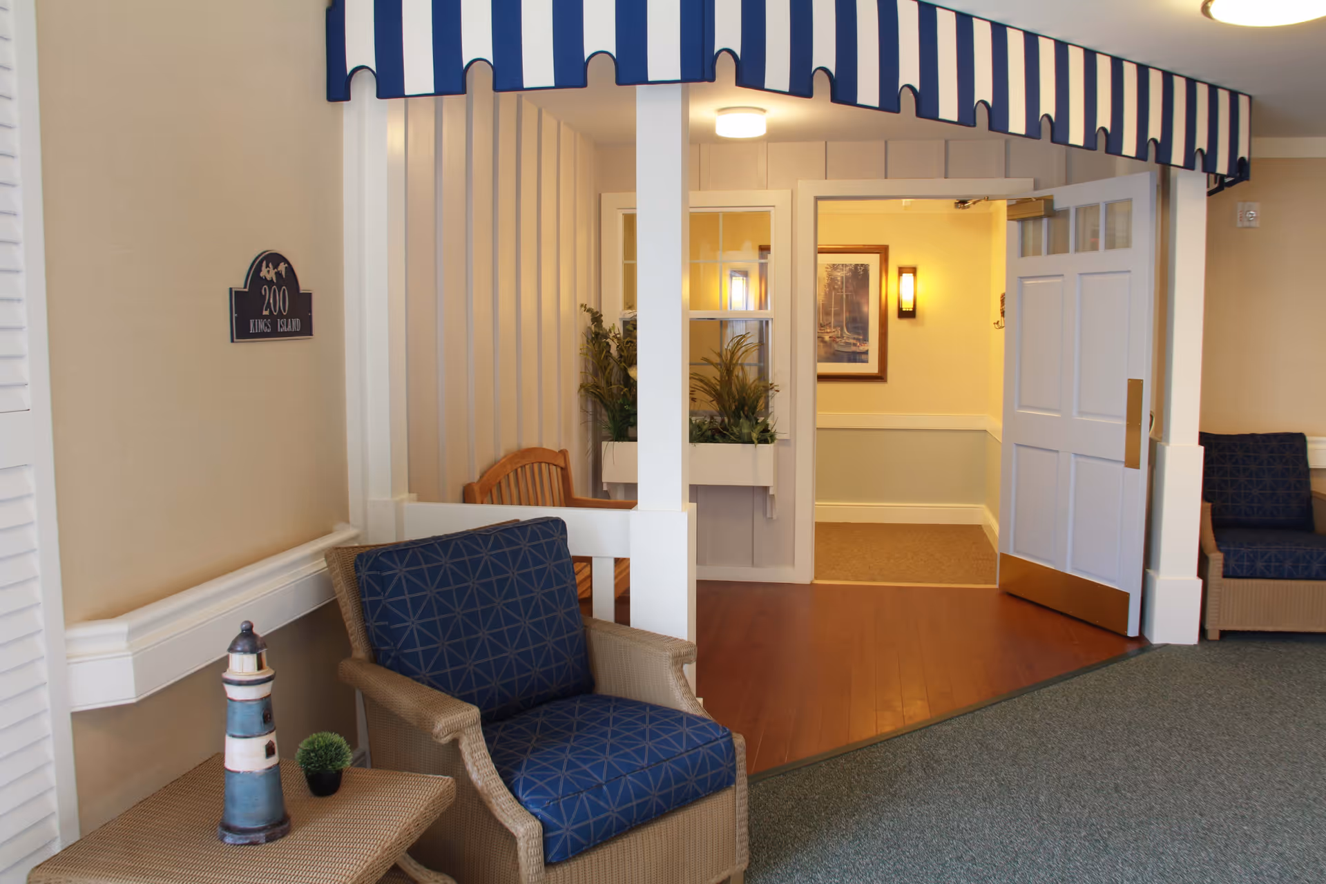Indoor seating area with two wicker chairs featuring blue patterned cushions, a small table with a lighthouse decoration and a small plant, a blue and white striped awning overhead, and an open door leading to a hallway with framed artwork and wall lighting.