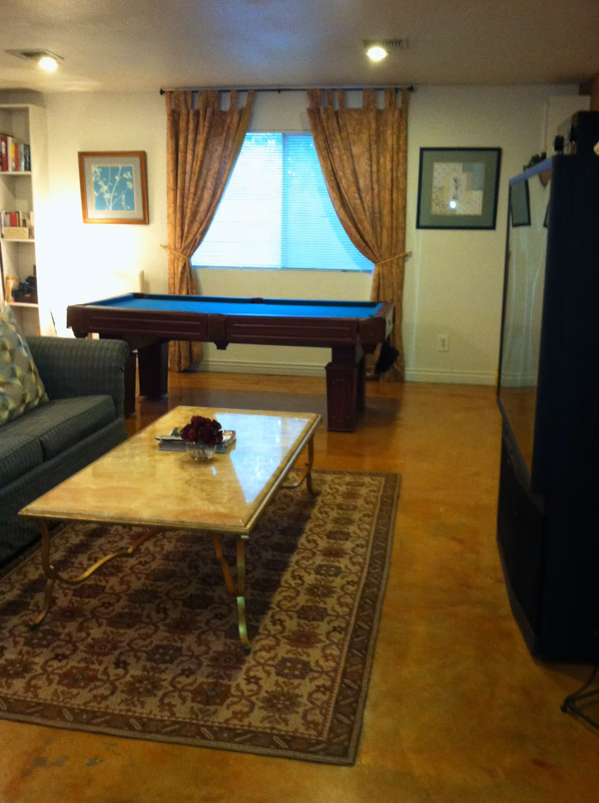 A cozy living room area with a patterned rug, a marble-top coffee table with a small flower arrangement, a blue upholstered couch with patterned pillows, a pool table near a window with gold curtains, framed artwork on the walls, and a large television on the right side.