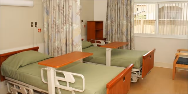 Two hospital-style beds with green bedding and overbed tables in a shared patient room with privacy curtains and a window.