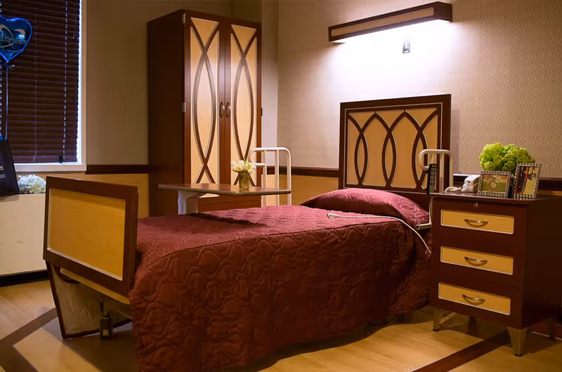 A neatly arranged nursing facility bedroom with a single bed covered in a burgundy quilt, bedside table, wardrobe, and over-bed table.