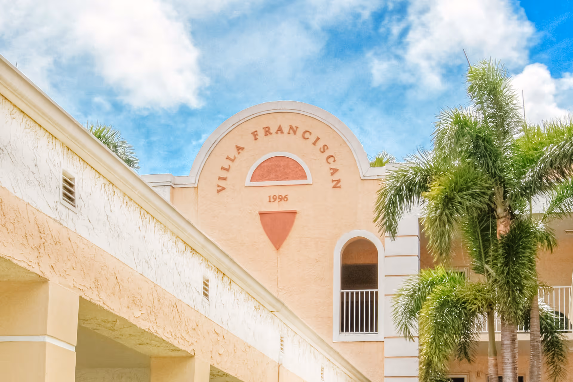 Exterior view of Villa Franciscan building with beige walls, arched windows, and palm trees under a partly cloudy blue sky.