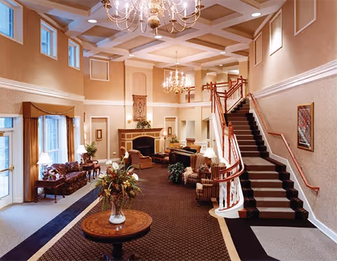 Spacious and elegant living room area with high ceilings, chandeliers, a staircase with wooden handrails, multiple seating arrangements including sofas and armchairs, a fireplace, large windows with curtains, and a round table with a floral arrangement in the center.