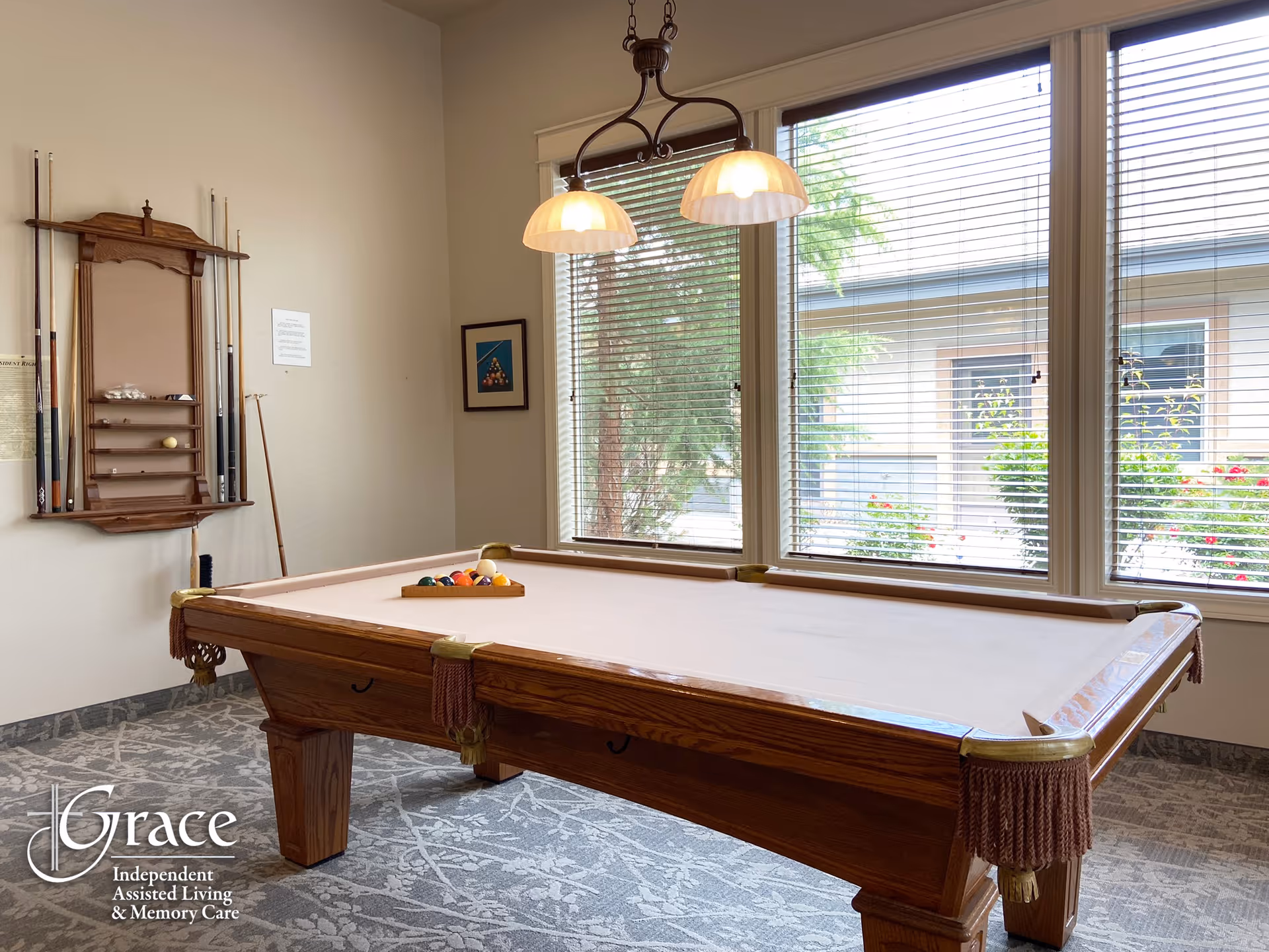 A well-lit room with a wooden pool table in the center, a rack with pool cues and balls mounted on the wall, large windows with blinds letting in natural light, and a hanging light fixture above the pool table. The carpet has a floral pattern, and there is a framed picture on the wall.