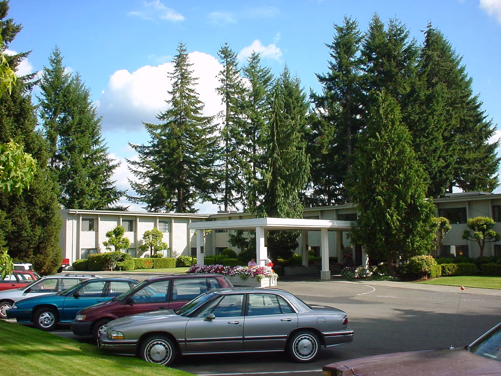Exterior view of Olympics West Senior Living facility showing a two-story building surrounded by tall evergreen trees, a covered entrance with white pillars, and several parked cars in the driveway under a partly cloudy blue sky.