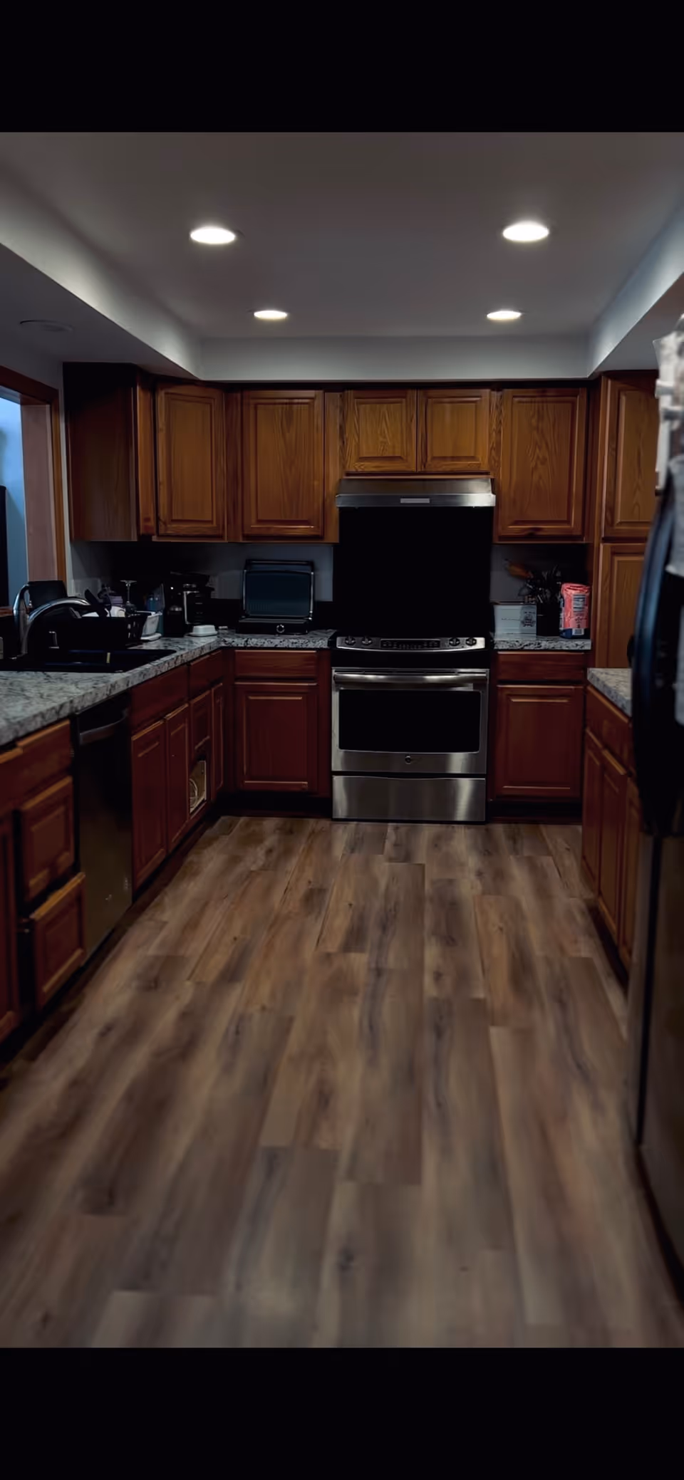 Galley-style kitchen with wooden cabinets, stainless-steel stove and dishwasher, granite countertops, and wood-look flooring under recessed ceiling lights.