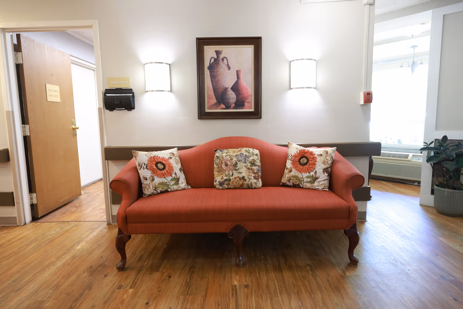 Red upholstered sofa with three decorative pillows beneath a framed painting in a bright seating area.