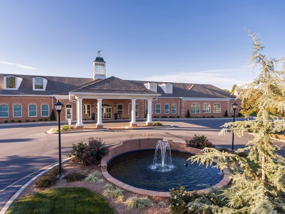 Exterior view of a single-story brick building with a covered entrance supported by white columns. In front of the building is a circular driveway with a round fountain in the center, surrounded by landscaping and bushes. The sky is clear and blue.
