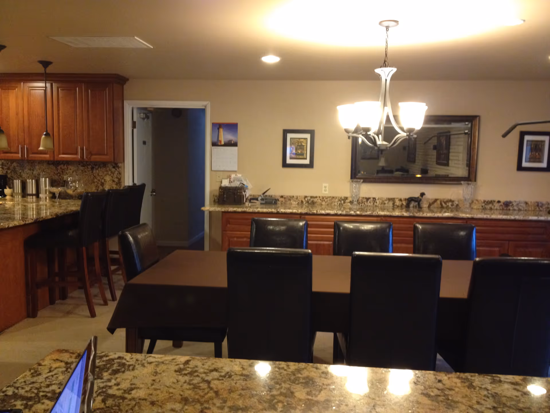 Interior view of a dining area with a long table covered with a brown tablecloth and surrounded by black chairs. Behind the table is a granite countertop with wooden cabinets underneath and a large mirror on the wall. To the left, there is a kitchen area with wooden cabinets, granite countertops, and bar stools. The room is lit by ceiling lights and a chandelier above the dining table.