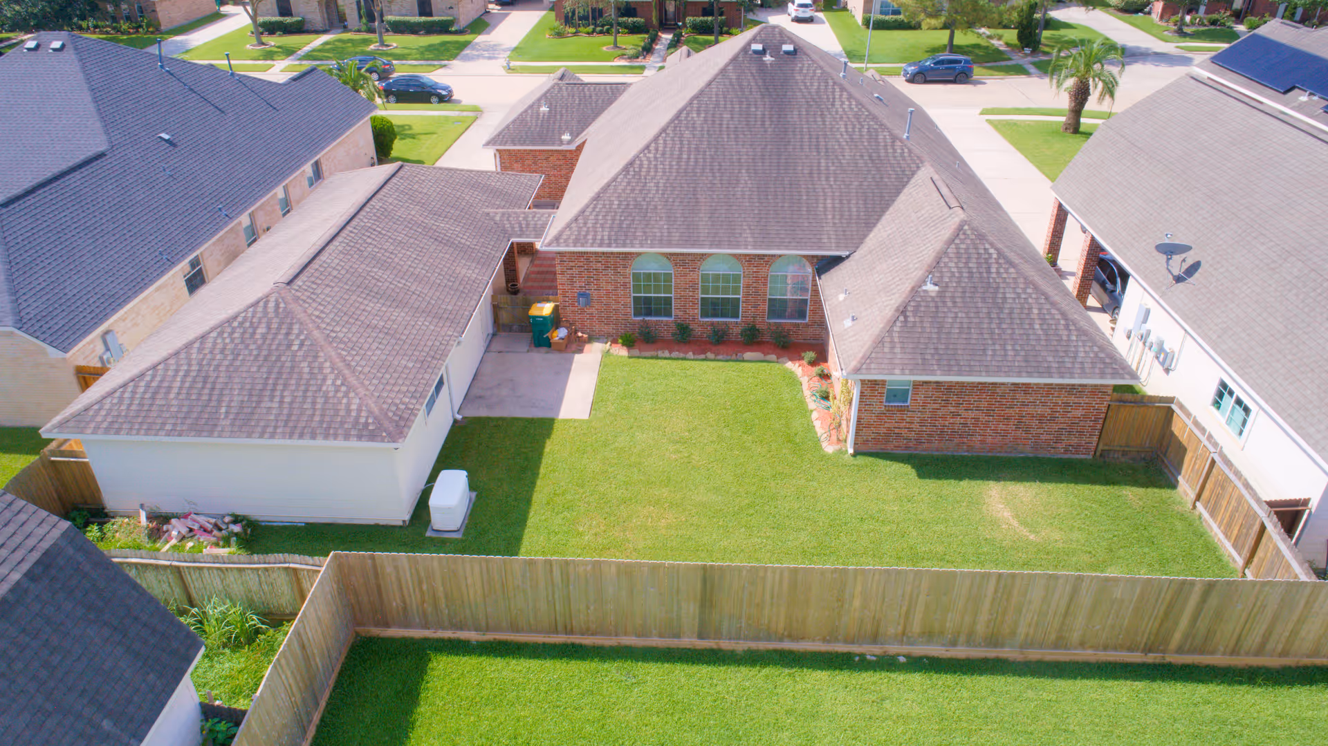 Aerial view of a residential backyard with a well-maintained lawn, surrounded by a wooden fence. The house has a brick exterior with multiple roof sections and several windows. Neighboring houses and a street with parked cars are visible in the background.