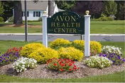 A green and white sign reading 'Avon Health Center' is displayed outdoors surrounded by colorful flower beds with red, white, purple, and yellow flowers. The background shows a grassy area and a residential-style building.