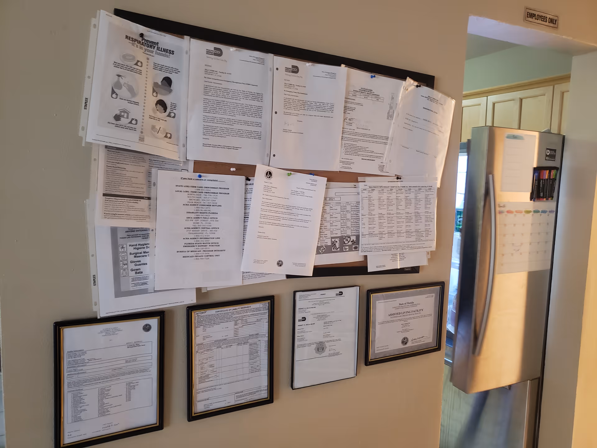 A bulletin board filled with various notices, documents, and schedules pinned on it, mounted on a beige wall. Below the bulletin board are four framed certificates or official documents. To the right, there is a stainless steel refrigerator with a calendar and some markers attached to it. Above the doorway next to the refrigerator is a sign that reads 'EMPLOYEES ONLY'.