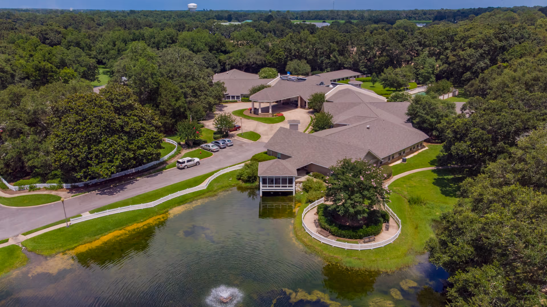 Aerial view of Live Oak Village senior living facility surrounded by lush green trees and a pond with a small fountain. The facility buildings have brown roofs and are arranged around a circular driveway with parked cars. There is a fenced circular garden area near the pond.