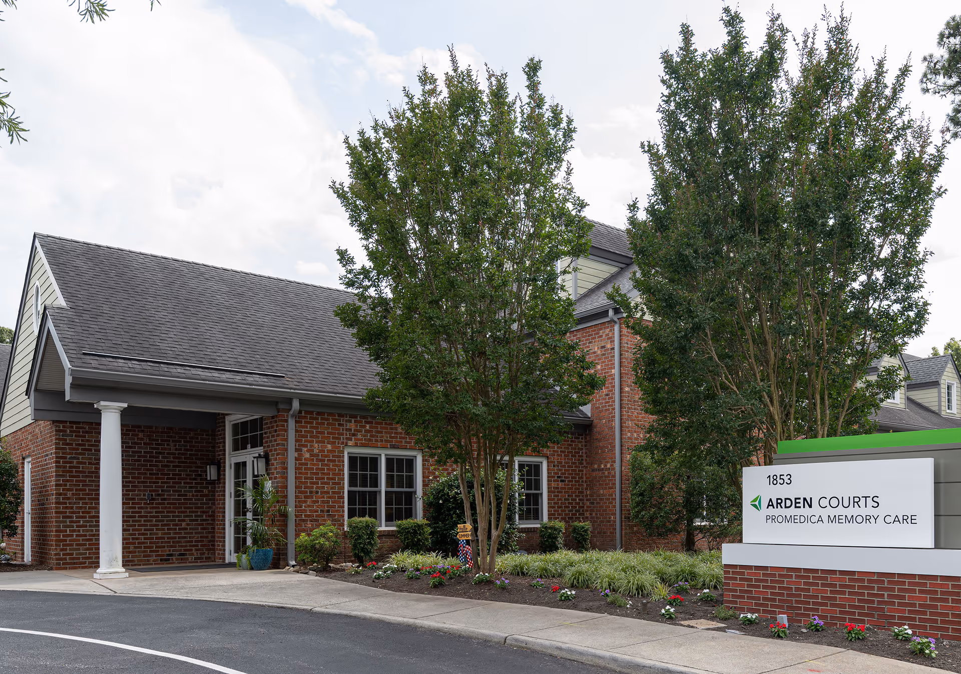 Exterior view of Arden Courts - ProMedica Memory Care Community building in Virginia Beach, showing a brick facade with white columns, windows, trees, and a landscaped garden area. A sign in front displays the facility name and address 1853.