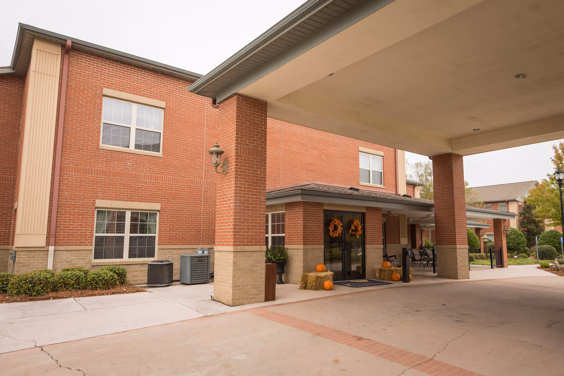 Entrance of a brick building with a covered driveway, decorated with autumn wreaths on the glass doors and pumpkins placed on hay bales near the entrance. The building has multiple windows and some greenery around the perimeter.