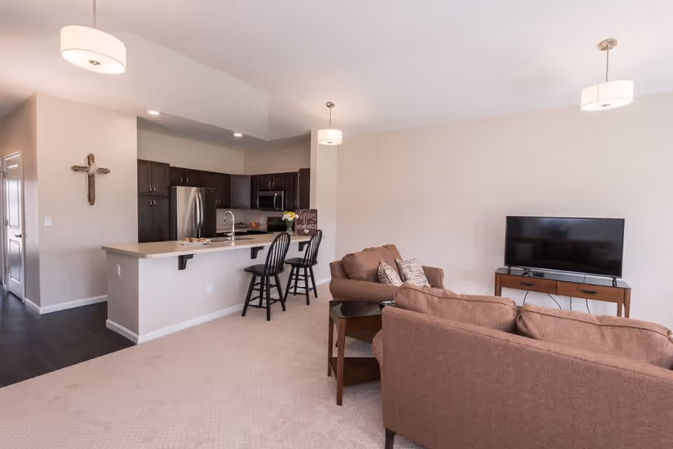 An open-concept living area featuring a kitchen with dark wood cabinets, stainless steel appliances, and a breakfast bar with two black bar stools. Adjacent to the kitchen is a living room with two brown sofas, a small side table, and a flat-screen TV on a wooden stand. The walls are light-colored, and there are three pendant lights hanging from the ceiling. A decorative cross is mounted on the wall near the kitchen entrance.