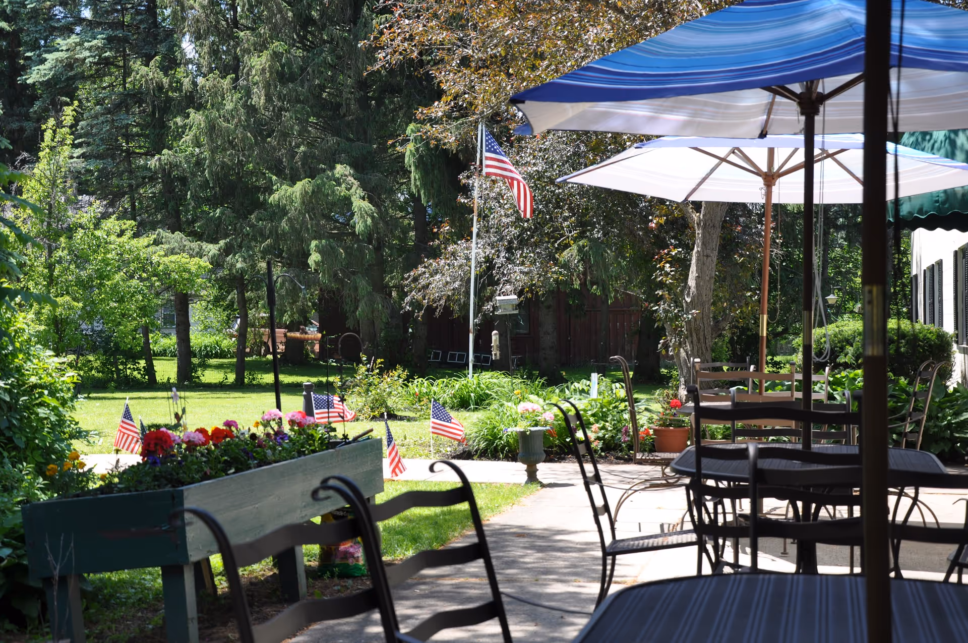 Outdoor patio area with metal tables and chairs under large umbrellas, surrounded by greenery, flowers, and small American flags. Tall trees and a flagpole with an American flag are visible in the background.