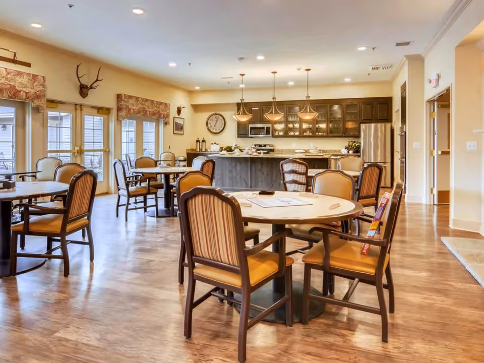 A spacious dining area in an assisted living community featuring round tables with cushioned chairs arranged on a wooden floor. The room has large windows with patterned valances, a kitchen area with hanging pendant lights, a clock on the wall, and stainless steel appliances including a refrigerator and microwave. Decorative elements include mounted antlers and framed pictures on the walls.