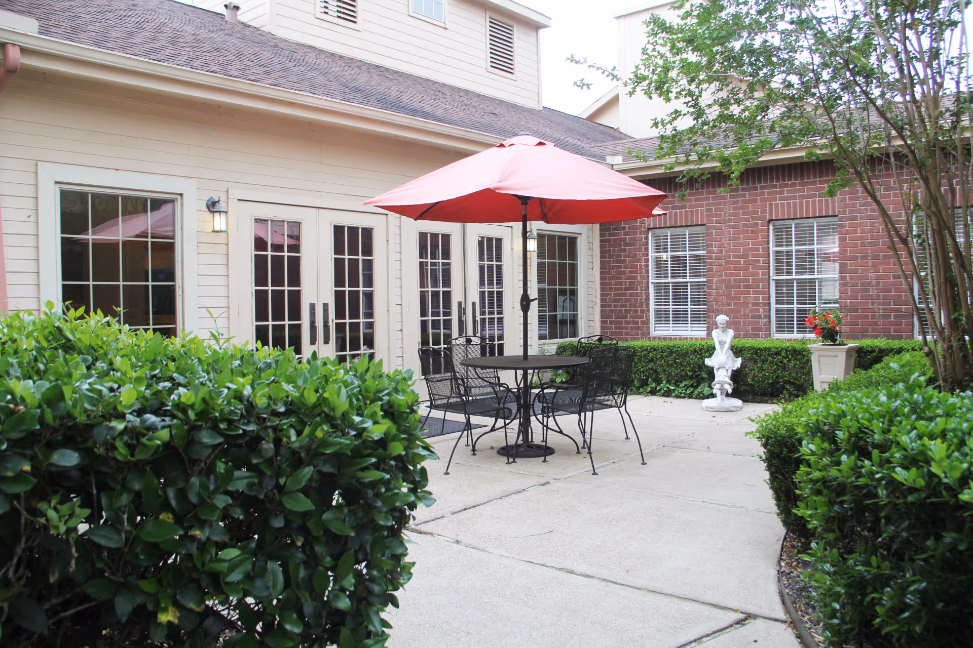 Outdoor patio area at Village on the Park Friendswood featuring a round black metal table with four matching chairs and a large red umbrella. The patio is surrounded by green bushes and trees, with a brick building and white-framed windows in the background. A small white statue and a planter with red flowers are also visible near the building.