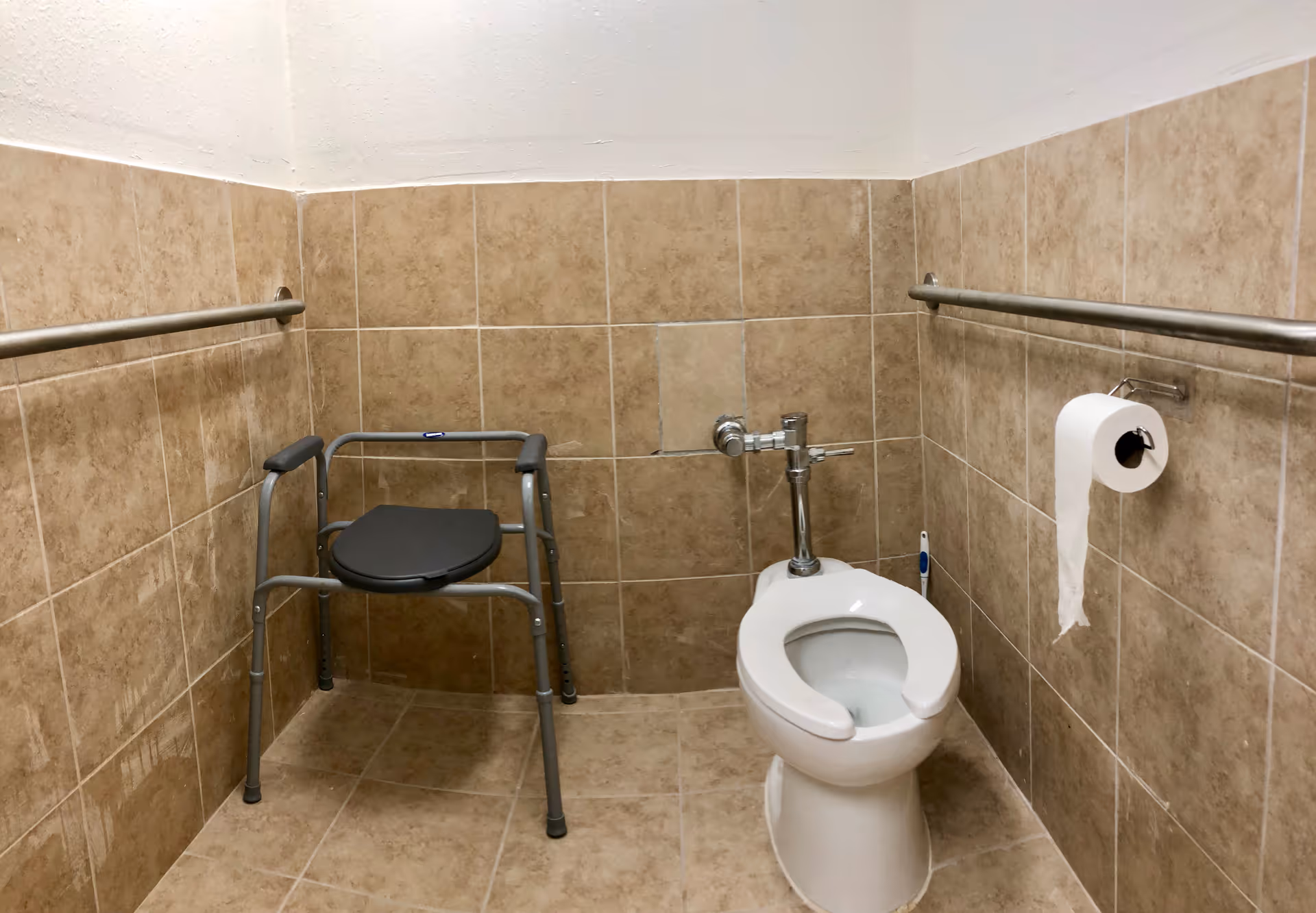 A small bathroom with beige tiled walls and floor, featuring a white toilet with an open front seat, a metal safety frame chair, stainless steel grab bars on both walls, and a toilet paper holder with a roll of toilet paper.