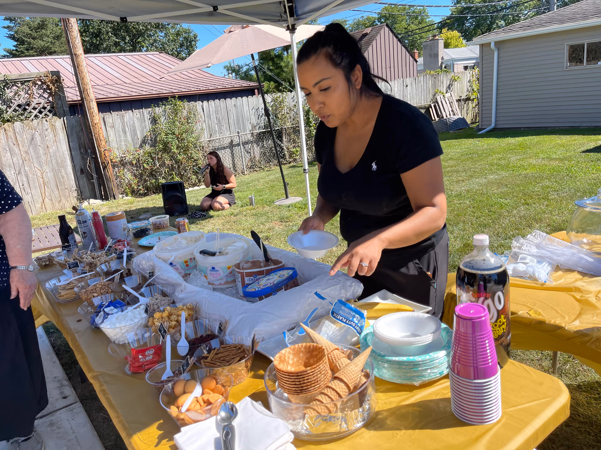 A woman is preparing a dessert table outdoors under a canopy. The table is covered with a yellow tablecloth and has various dessert ingredients including ice cream, cones, pretzels, marshmallows, and soda bottles. In the background, a young woman is sitting on the grass near a wooden fence and a small building.