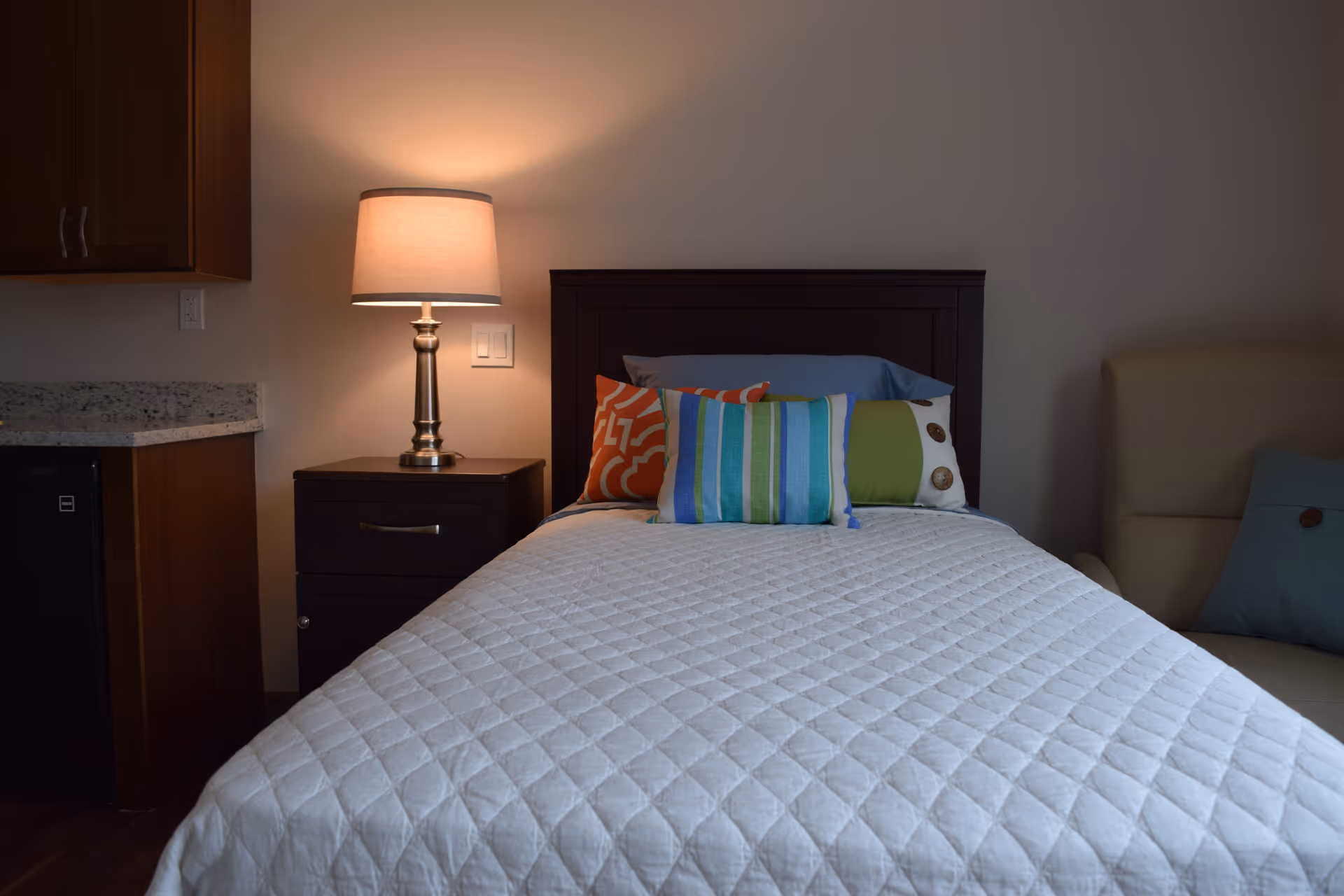 A neatly made bed with a white quilt and colorful pillows, a bedside table with a lit lamp, and parts of a kitchenette and armchair in a cozy bedroom.