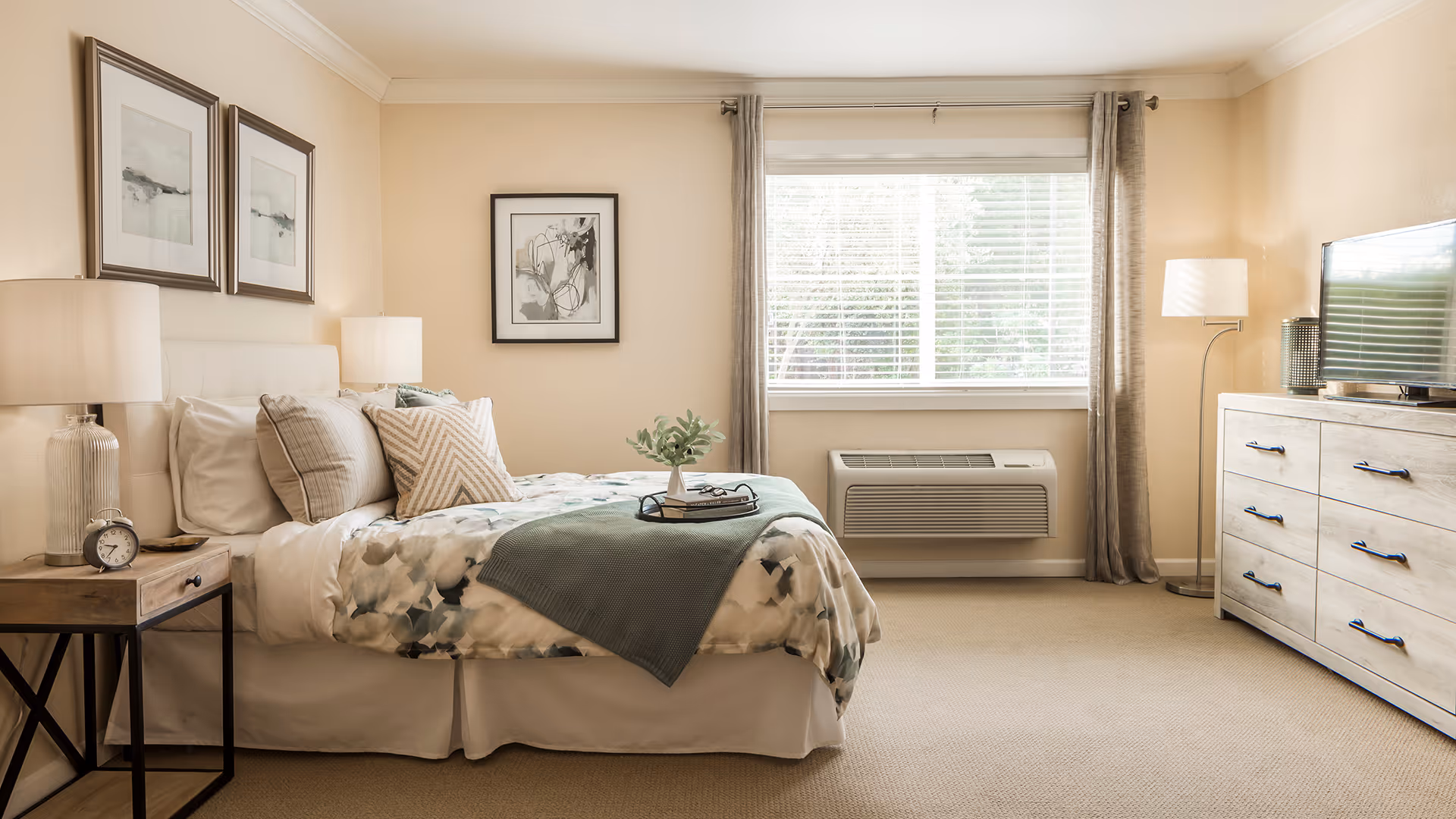 A bright and cozy bedroom with a neatly made bed featuring floral bedding and multiple pillows. There are two framed artworks above the bed, a wooden nightstand with a lamp and an alarm clock, a large window with curtains, a floor lamp, a dresser with a TV on top, and beige carpeting.