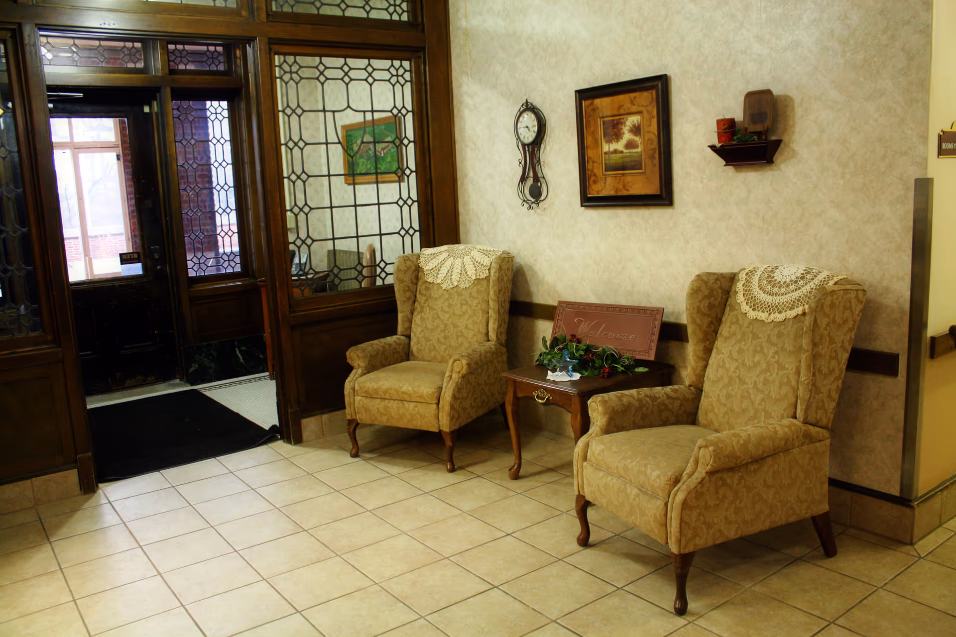 A cozy seating area in a senior living facility with two beige patterned armchairs featuring lace doilies on the headrests. Between the chairs is a small wooden table with a floral arrangement and a welcome sign. The walls are decorated with a framed picture and a vintage-style clock. The floor is tiled, and there is a wooden-framed glass door and window leading to another room.