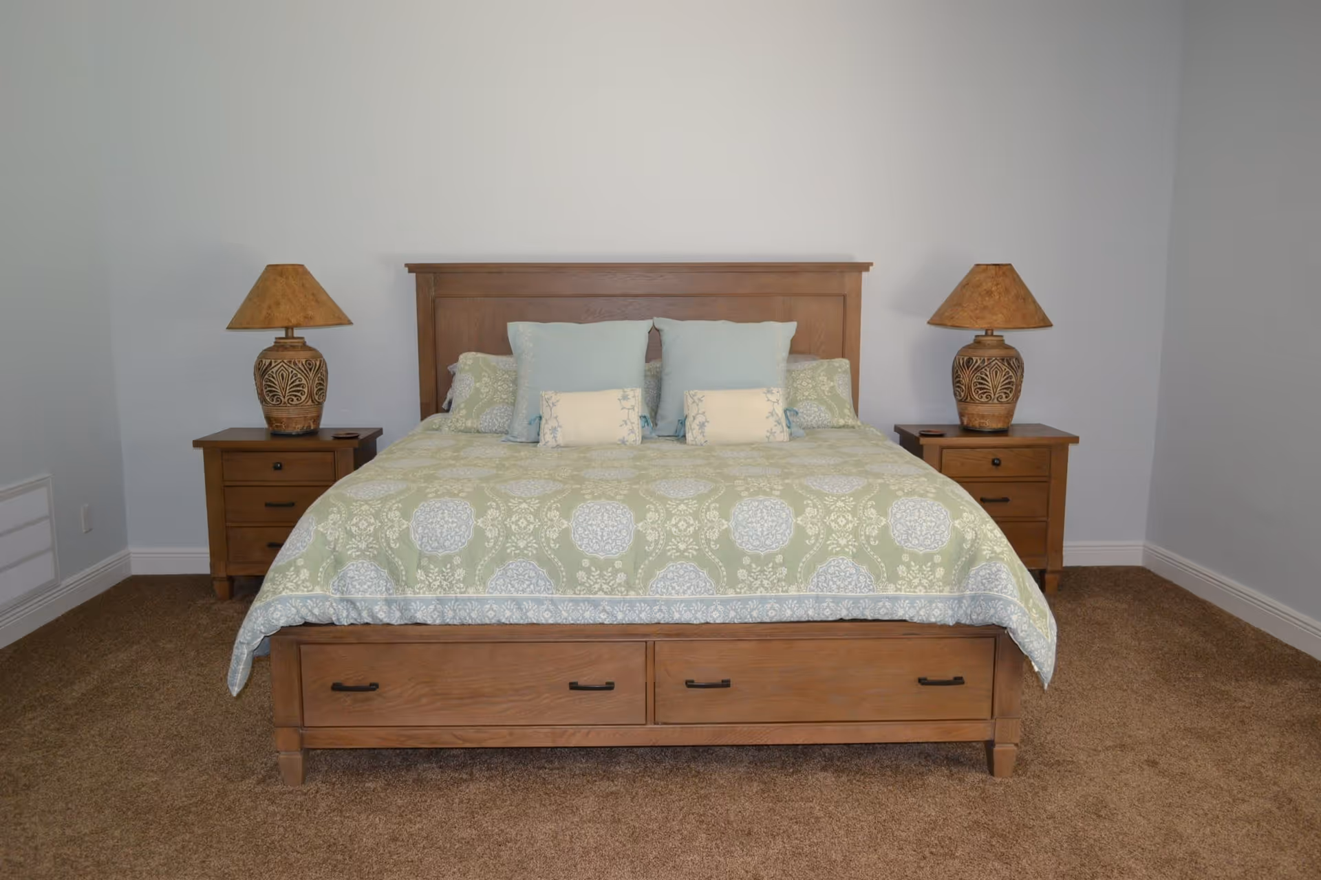 A bedroom with a wooden bed frame featuring two drawers at the foot of the bed. The bed is made with a green and blue patterned bedspread and has four pillows arranged at the head. On each side of the bed, there is a wooden nightstand with a decorative lamp. The room has beige carpet and plain white walls.
