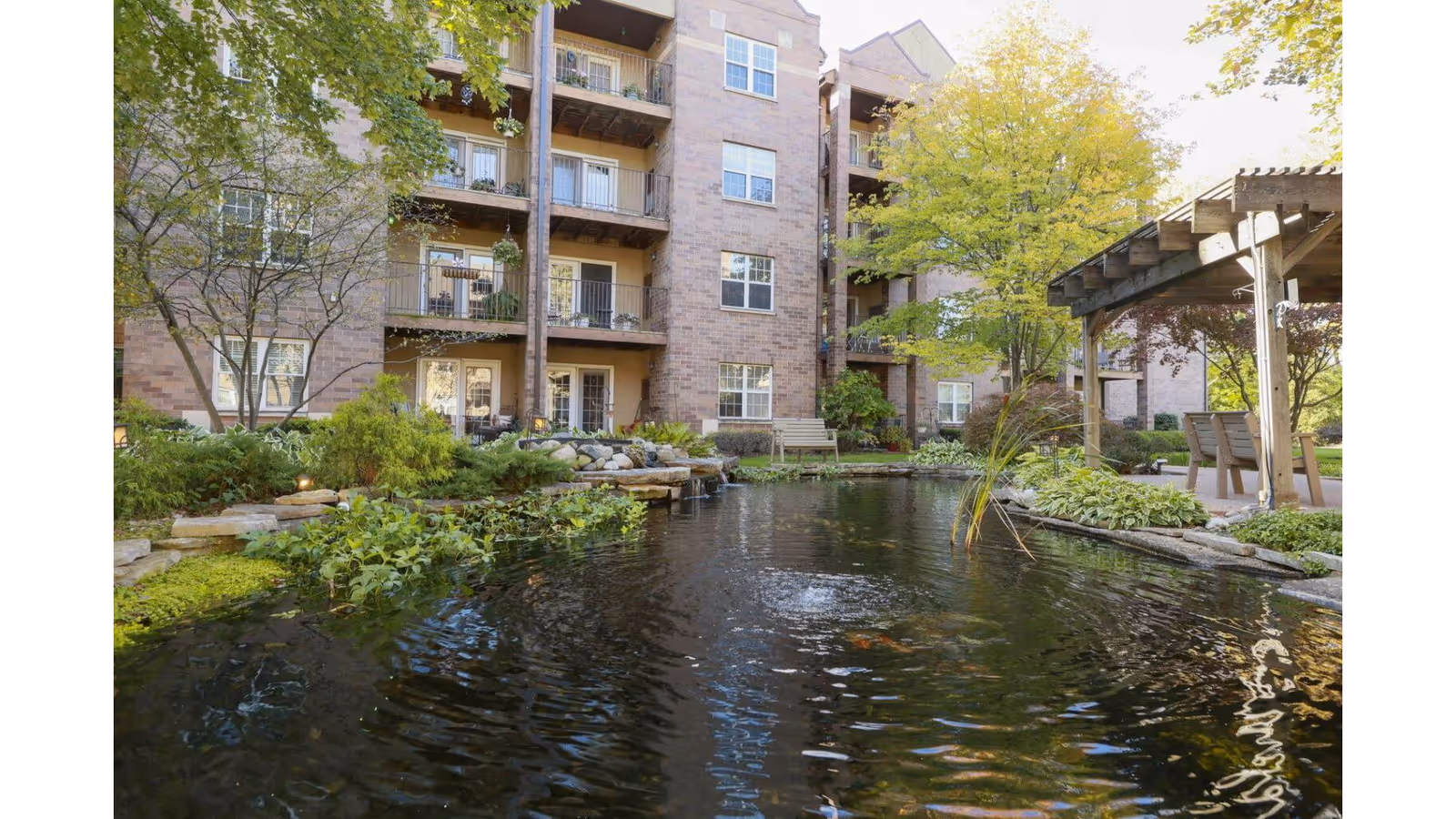 A landscaped courtyard with a koi pond, pergola, benches, and a multi-story brick residential building in the background.