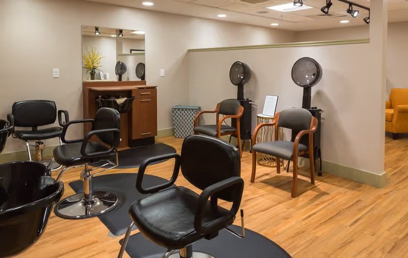 Interior view of a hair salon area with black salon chairs, hair washing stations, and hair dryers. The room has wooden flooring, beige walls, and a mirror above a wooden cabinet with a vase of yellow flowers.