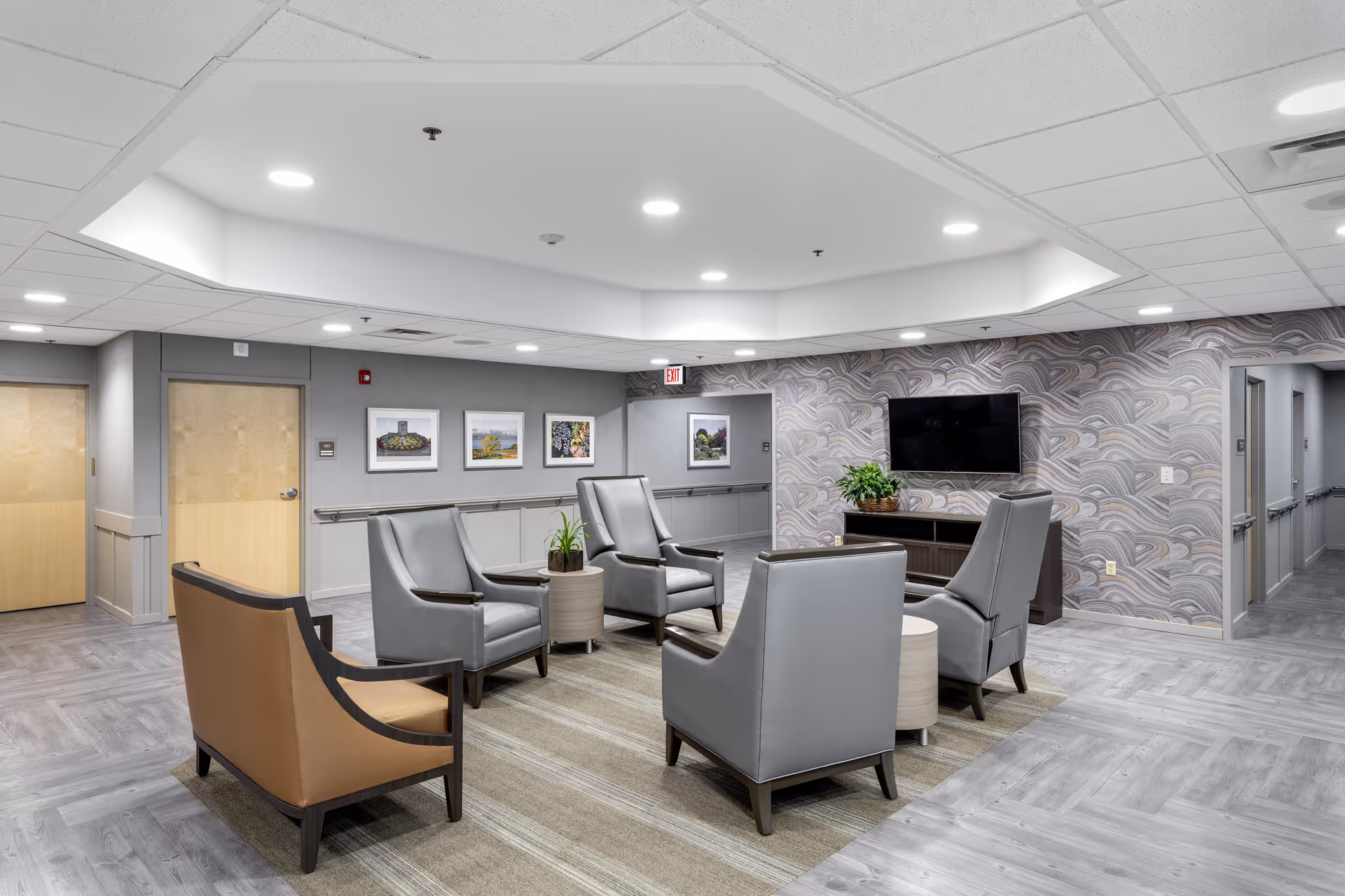 A modern senior living facility common area with five armchairs arranged around two small round tables on a beige rug. The walls are decorated with framed pictures and a flat-screen TV mounted on a patterned accent wall. The ceiling has recessed lighting and the floor is covered with light gray wood-like flooring.