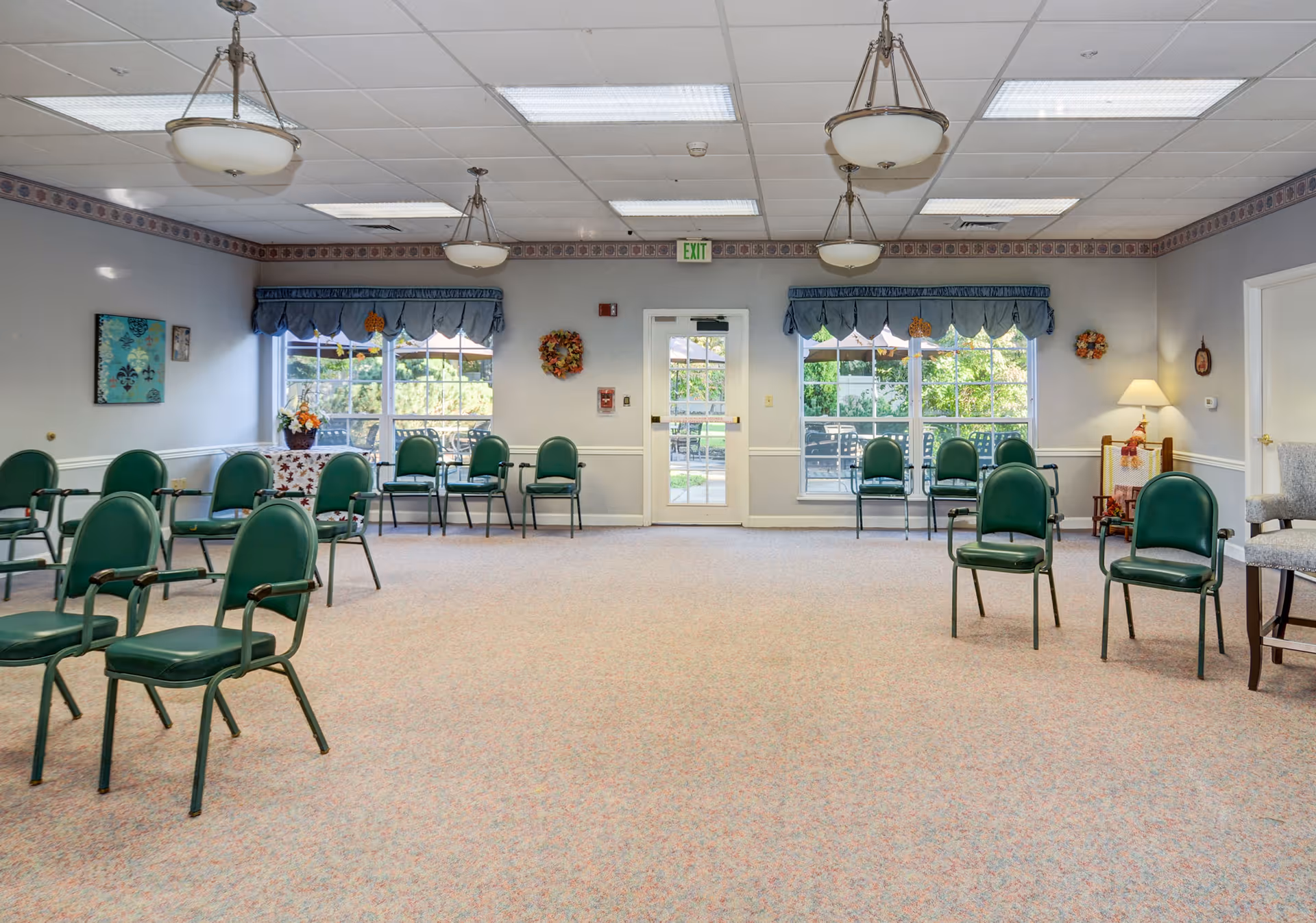 A spacious room with multiple green chairs arranged in a semi-circle and along the walls. The room has large windows with blue valances, allowing natural light to enter. There are three ceiling light fixtures, a door in the center of the far wall, and some decorative wreaths and framed artwork on the walls. The carpet is light-colored with a subtle pattern.