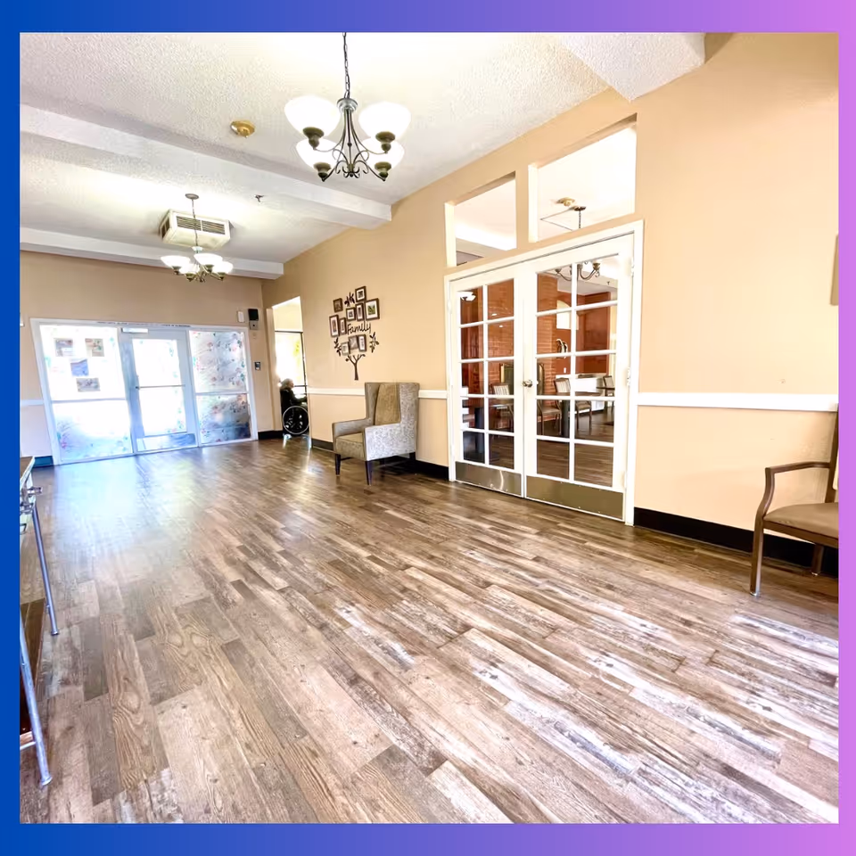 Bright senior living facility lobby with wood-look flooring, chairs, chandeliers, and glass double doors.